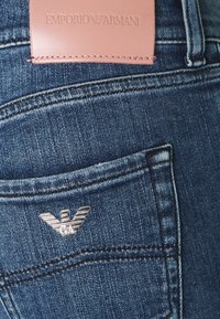 Mörkblå denimjeans med en läderpatch med "Emporio Armani"-logotyp och en silverfärgad metall-logotyp på bakfickan. Slät textur.