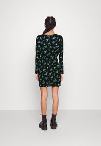 Robe noire à manches longues ornée de motifs floraux bleus et verts, taille cintrée et fermeture à bouton dans le dos. Associée à des bottes noires à talon chunky.