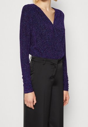 Kobieta nosząca długi rękaw, głęboko fioletowy, błyszczący top kopertowy wpuszczony w czarne szerokie spodnie z wysokim stanem.