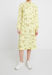 Donna che indossa un vestito lungo fino al ginocchio, a maniche lunghe, di un giallo chiaro con piccoli motivi floreali marroni, abbinato a delle sneakers bianche.
