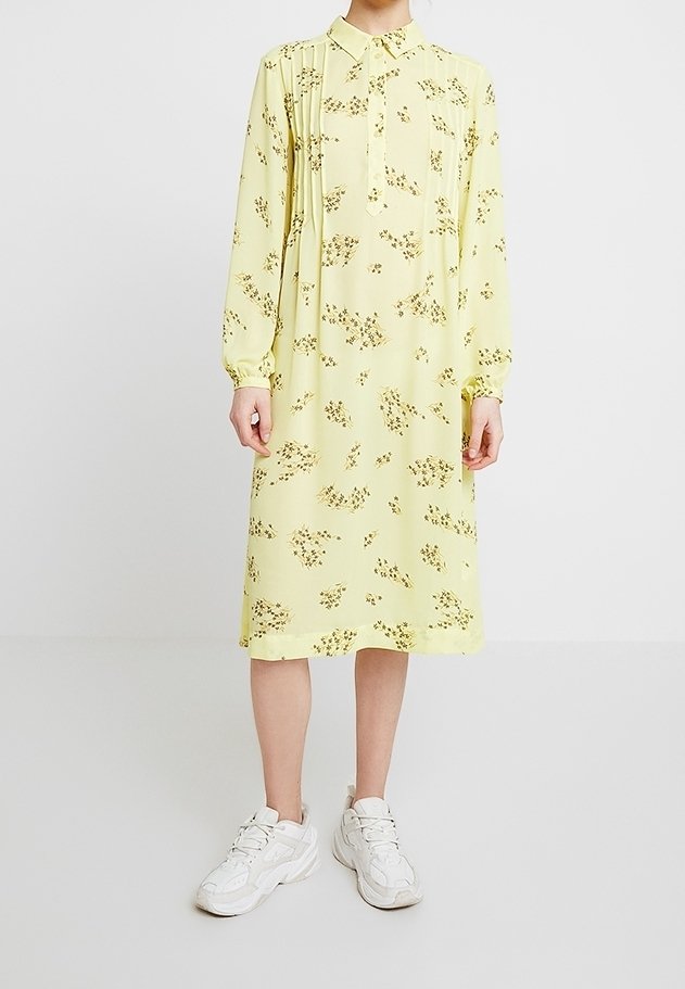 Donna che indossa un vestito lungo fino al ginocchio, a maniche lunghe, di un giallo chiaro con piccoli motivi floreali marroni, abbinato a delle sneakers bianche.