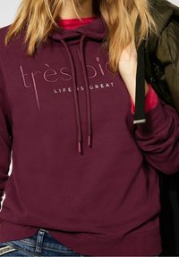 Bordowa bluza z kapturem z teksturowanym logo z napisem „très bien” i „LIFE IS GREAT.” Posiada sznurki regulacyjne i miękkie wykończenie materiału.