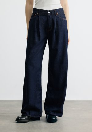 Person iført vidde-ben mørkeblå denimbukser, hvid top og sorte sko, stående på en grå gulv mod en ensfarvet hvid baggrund.