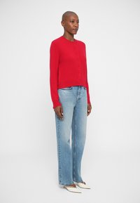 Červený kardigán s dlhým rukávom, okrúhlym výstrihom a zapínaním na gombíky, v kombinácii s svetlomodrými džínsy s vysokým pásom a bielymi lodičkami so špicatou špičkou.