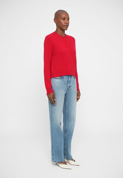 Cardigan rouge à manches longues avec encolure ronde et fermeture à boutons, associé à un jean taille haute bleu clair et des chaussures blanches à bouts pointus.