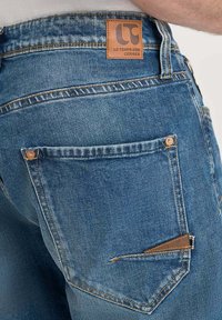 Jeans en denim bleu clair avec une patch en cuir marron à l'arrière. Le design comprend une seule poche arrière avec des détails de couture et des rivets.