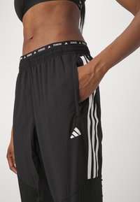 Czarne sportowe spodnie z elastycznym pasem, bocznymi kieszeniami i białymi pionowymi paskami. Pas i dół spodni ozdobione logo.