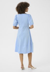 Svetlo modra bombažna obleka z napihnjenimi rokavi, dolžine do kolen in brezšivnim hrbtom. Obuta z bež slingback čevlji. Minimalističen dizajn.