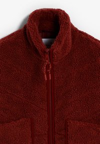 Chaqueta de forro polar roja con cuello alto, cremallera completa y tejido texturizado con costuras visibles y dos bolsillos frontales.
