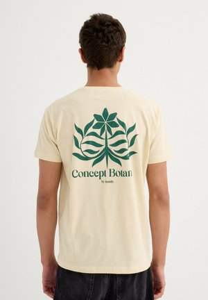 Camiseta de algodón beige con un estampado botánico verde en la parte posterior, que presenta flores y hojas estilizadas, y texto que dice "Concept Botan by inside."