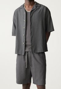 Camisa de manga corta gris oscuro con cierre de botones y cuello, combinada con pantalones cortos gris a juego. Ambas prendas tienen un corte relajado.