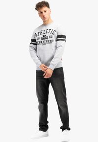 Felpa grigia con stampa grafica nera, caratterizzata da due strisce nere orizzontali sulle maniche. Abbinata a jeans scuri e sneakers bianche.