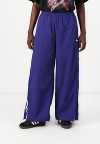 Lilla joggebukser med elastisk midje, stor løs passform og hvit Adidas-logo på siden. Har glidelås på sidene.