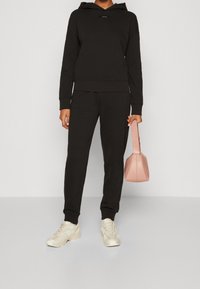 Set di felpa nera e pantaloni da tuta realizzati in tessuto morbido, abbinati a sneakers beige. Tiene in mano una piccola borsa rosa con un design strutturato.