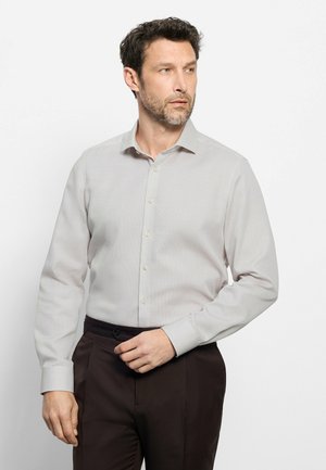 Man met donker haar en baard, gekleed in een lichtgrijs, langemouwig overhemd met knopen en een donkere broek, kijkt iets omhoog naar de zijkant.