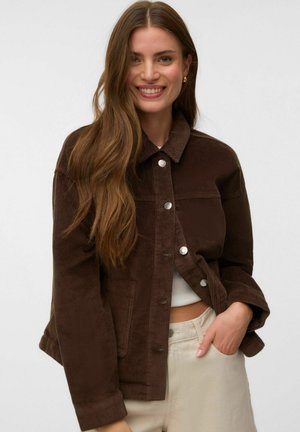 Femme souriante aux longs cheveux bruns porte une veste en velours côtelé marron foncé et un pantalon clair, une main dans la poche.
