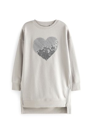 Barn iført en lysegrå lang sweatshirt med et sølvglitrende hjerte foran og blå jeans, står med armene afslappet ved siden.