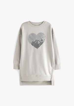 Barn iført en lysegrå lang sweatshirt med et sølvglitrende hjerte foran og blå jeans, står med armene afslappet ved siden.