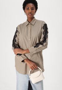 Cămașă bej cu dungi, cu mâneci din dantelă neagră, închidere cu nasturi și guler deschis. Modelul ține o mică poșetă albă. Fusta din denim este vizibilă.