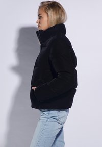 Sort puffjakke med høj krave, frontlynlås og sidelommer. Blød tekstur, cropped design, parret med lyseblå jeans.