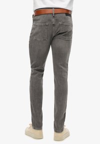 Jeans de mezclilla gris con un ajuste slim. Presenta cinco bolsillos y un cinturón marrón. La tela tiene una textura suave y los puños son ligeramente ajustados.