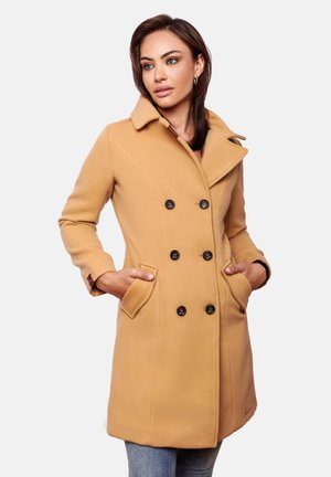 Vrouw draagt een dubbelrijige jas in camel kleur met zwarte knopen, handen in de zakken, kijkt iets naar de zijkant tegen een effen achtergrond.