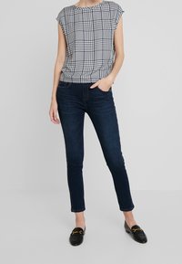 Donna che indossa un top senza maniche a quadretti bianco e nero, jeans skinny blu scuro e mocassini neri con accessori dorati.