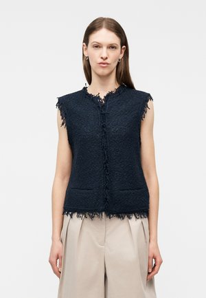 Woman wearing sleeveless textured black vest with fringed edges and beige pleated pants, standing against a plain light background.