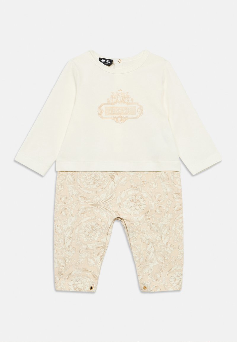 Λευκή μπλούζα με μακριά μανίκια και ανάγλυφο χρυσό λογότυπο· μπεζ παντελόνια με μοτίβο και floral σχεδιασμό, με χρυσά κουμπώματα στα μπατζάκια.