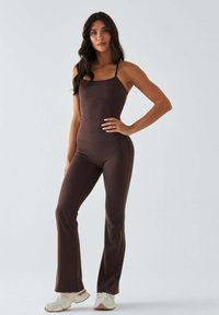 Brązowy dopasowany kombinezon na cienkich ramiączkach, z rozszerzanymi nogawkami i gładką fakturą. Połączony z jasnymi butami sportowymi. Przezroczyste tło.