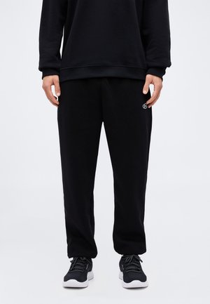 Mann trägt schwarzen Sweatshirt, schwarze Jogginghose mit kleinem weißem Logo am linken Oberschenkel und schwarze Sneakers mit weißen Sohlen, steht vor weißem Hintergrund.