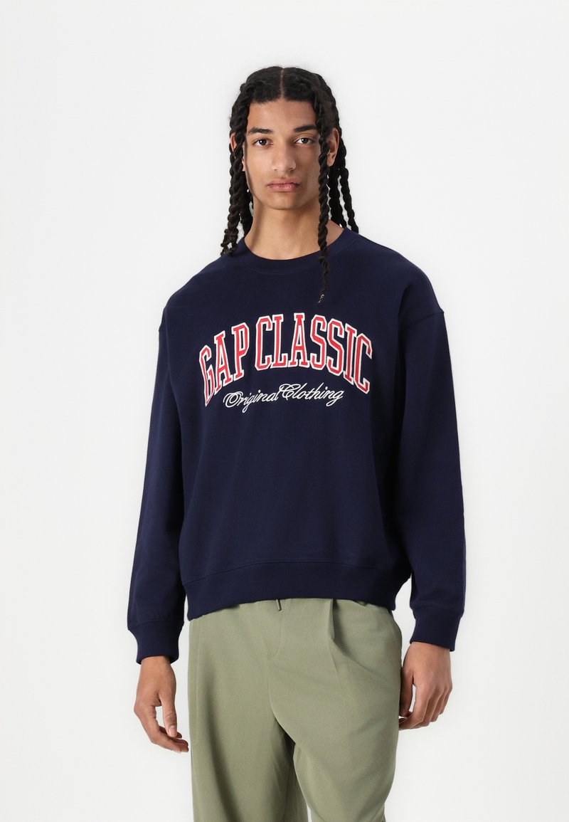 Mörkblå sweatshirt med texten "GAP CLASSIC" och "Original Clothing" i röd och vit. Har en rundad halsringning och ribbade ärmslut.