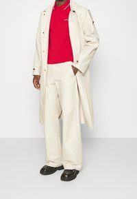 Beige trenchcoat över en röd polo med en logotyp, tillsammans med vita byxor och svarta plattformsskor. Slät tygtextur, avslappnad passform och minimal detaljering.