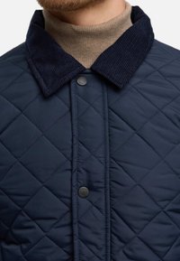 Chaqueta acolchada azul marino con cuello de pana, cuenta con un cierre frontal de botones, patrón de diamantes y textura suave.
