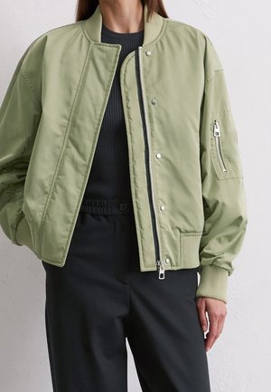Person wearing light green bomber jacket with zipped pocket on sleeve, black ribbed top, and black high-waisted pants against white background.