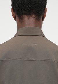 Πουκάμισο με γιακά σε απαλό γκρι-καφέ χρώμα, με ραμμένη ραφή στο πίσω μέρος και δύο διακριτικές επωνυμίες κειμένου.