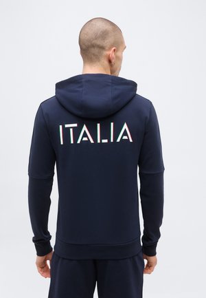 Mees paljaste juuste ja tumesinise kapuutsiga, millel on taga kirjas "ITALIA" punastes, valgetes ja rohelises kirjades, seisab ühetaustalise heleda tausta ees.