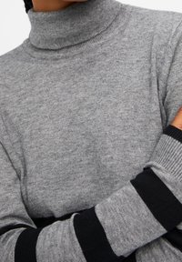 Pull à col roulé gris en tissu tricoté doux, avec des poignets rayés noirs et gris. Vue rapprochée mettant en valeur la texture et le design.