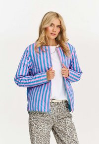 Puchowa kurtka z pionowymi niebieskimi i różowymi paskami, pikowanym wzorem, zamek z przodu oraz elastycznymi mankietami. W zestawie z beżowymi spodniami w cętkowany wzór lamparta.