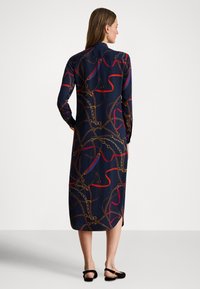 Γυναίκα με πλάτη, φοράει midi φόρεμα σε ναυτικό μπλε χρώμα με σχέδιο αλυσίδων και σχοινιών, μακριά μανίκια, σε συνδυασμό με μαύρες επίπεδες γόβες με λουράκι στο πίσω μέρος.