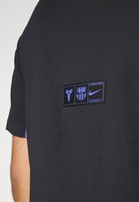 Zwart T-shirt met een opvallende patch met logo's in het blauw, inclusief een sportlogo en tekst. Korte mouwen met blauwe accenten. Zachte textuur.