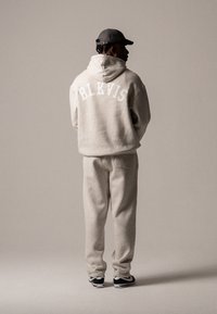Beiger Pullover mit weißer "BLKVIS" Schriftzug, kombiniert mit passenden grauen Jogginghosen und schwarzen Sneakers, von hinten betrachtet.