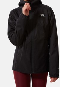 Person, der eine schwarze Regenjacke von The North Face zuknöpft, trägt dunkelrote Leggings, von den Schultern bis zu den Oberschenkeln zu sehen.