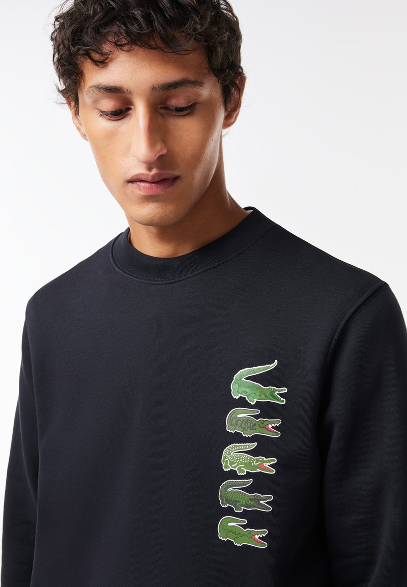 Sudadera negra con una fila vertical de gráficos de cocodrilo verdes en el lado izquierdo, mostrando diseños y expresiones variadas.