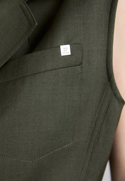 Tamsiai žalia be rankovių apranga su kišene ir sidabriniu logotipo akcentu bei tekstūruotu audiniu. Matomi tvarkingi siuvimo detalės.