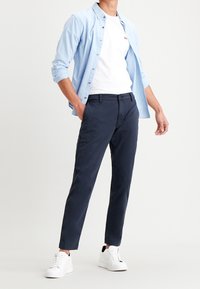 Chemise boutonnée bleu clair portée sur un T-shirt blanc, associée à un pantalon chino bleu marine et des baskets blanches. Style décontracté, coupe ajustée avec des accents simples.