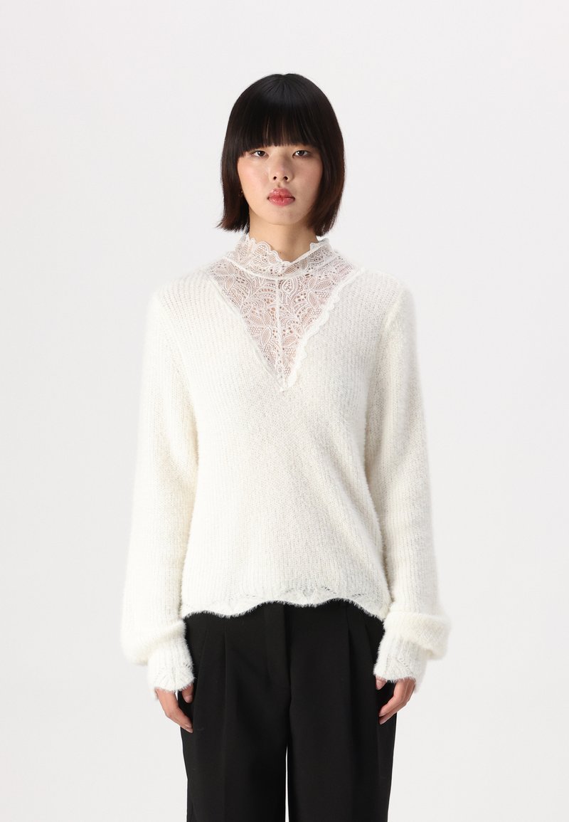 Pull blanc en tissu doux et duveteux, avec un panneau en dentelle au niveau du col et des manches larges, associé à un pantalon noir.