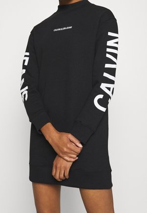Person trägt ein langes schwarzes Calvin Klein Sweatshirtkleid mit weißem Logo auf der Brust und großem Text auf beiden Ärmeln.