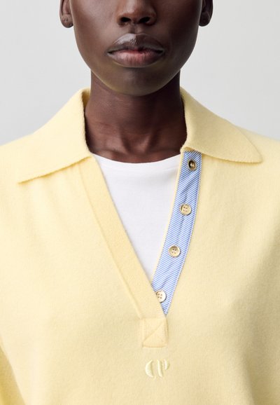 Suéter amarillo claro con escote en V y una tapeta a rayas azul en contraste. Presenta botones decorativos y un sutil detalle de logo.