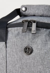 Γκρι υφασμάτινος σακίδιο με υφή, που διαθέτει μαύρους κωδικούς τσέπης, μια διακοσμητική κουμπιά και ανθεκτικό λουρί μεταφοράς.